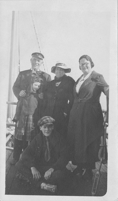 Black-and-white photograph of five people on the deck of a ship. They are standing against a railing, and rigging can be seen in the background Elizabeth Foss Wilson and Helen Wilson Williams stand next to an older man and a young girl. Edward Foss Wilson is seated at their feet on the ship deck. All are white people. All are wearing coats. All but the little girl are smiling into the camera.