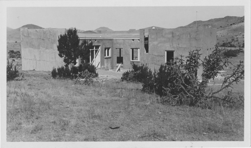 Black-and-white photograph of a partially-built stucco building. Scrub-brush surrounds the building, and desert mountains are in the background.