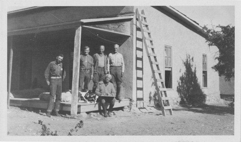 Black-and-white photograph of five white men standing on edge of covered porch of stucco building. Three men - including Thomas E. Wilson - are standing on the porch, one is standing just off of the porch, and one is seated on the edge of the porch. All are wearing work clothes.