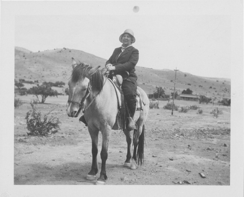 Black-and-white photograph of Elizabeth Foss Wilson seated on a light-colored horse in New Mexico desert. Scrub-brush, a few low-lying buildings, and desert mountains are behind her. Elizabeth is an older white woman wearing a dark jacket and skirt, and a light-colored cloche hat.