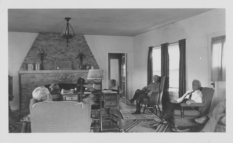 Black-and-white photograph of three white women and four white men seated in living room of house. House is sparsely furnished, and has a central stone fireplace.