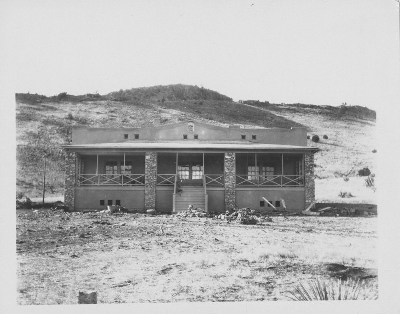 Black-and-white photograph of one-story stucco building with wide front porch and central staircase. Desert hills are in the background.