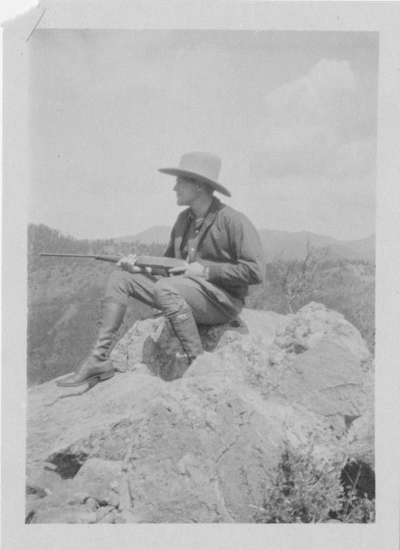 Black-and-white photograph of young Edward Foss Wilson seated on a rocky mountaintop. He is wearing a cowboy hat and calf-length boots, and is resting a rifle across his lap. He is photographed in three-quarters profile. Mountains are pictured in the background.