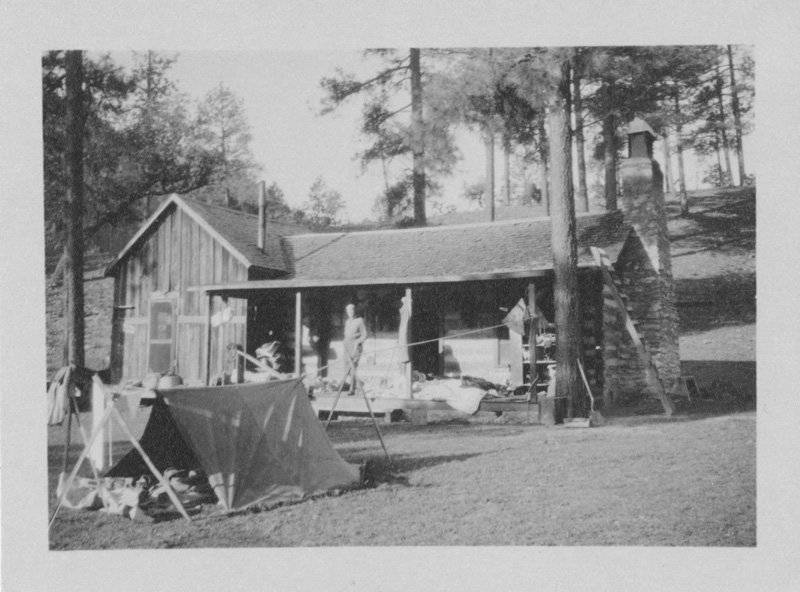 Black-and-white photograph of wooden cabin with stone chimney surrounded by pine trees. A white man stands on the porch. A tent is pitched in the yard.