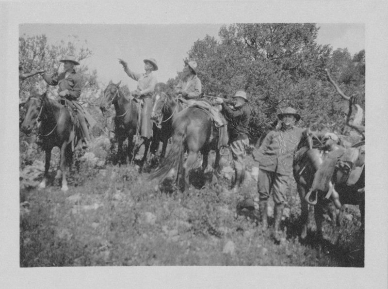 Black-and-white photograph of five white men and five horses on a grassy hill with pine trees behind them. Three men are riding their horses, and two are standing next to their horses. Thomas E. Wilson is pictured standing next to his horse, and he is aiming a rifle across the horse's back to a point off-camera. One of the seated riders is pointing in the same direction that Thomas E. Wilson is aiming his gun.