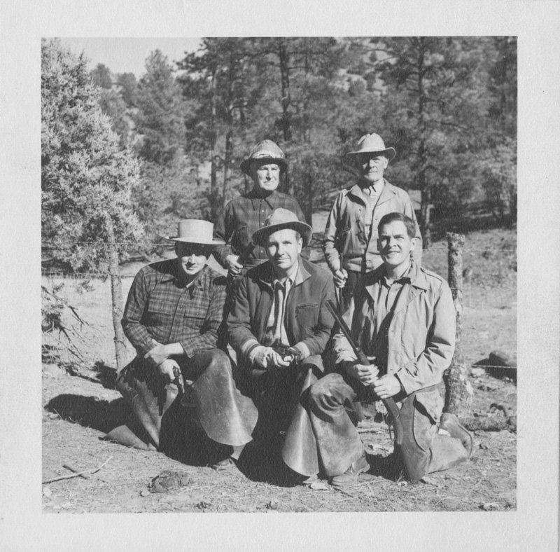 Black-and-white photograph of five white men wearing chaps and cowboy hats and holding guns. Three are kneeling, and the other two are standing behind them. Pine trees are shown behind them.