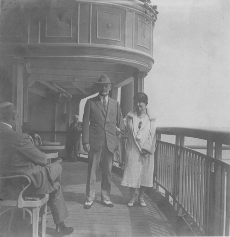 Black-and-white photograph of Thomas E. Wilson and Elizabeth Foss Wilson standing on a balcony. Both are white and middle-aged. Thomas wears a light colored suit and hat, and Elizabeth wears a white fur-trimmed coat and dark cloche hat.