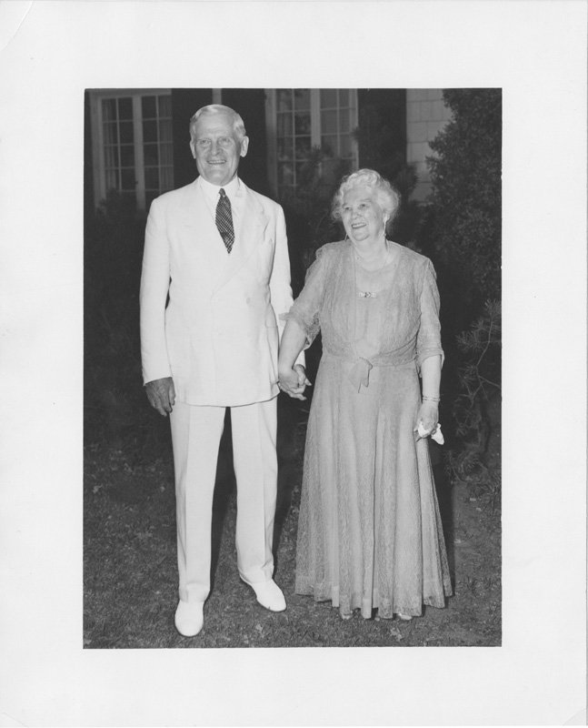 Black-and-white photograph of Thomas E. Wilson and Elizabeth Foss Wilson standing outdoors on a lawn. They are a white man and white woman with white hair, shown smiling and holding hands. Thomas is wearing a white suit and Elizabeth is wearing a long light-colored gown.