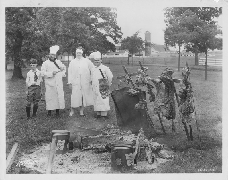 Black-and-white photograph of two white men and one white woman wearing chef's clothing, and a young white boy, standing around an open fire pit with spits of roasting meat. The fire pit is in a grassy field. Behind it are trees, a fence, and distant farm buildings. Boy appears to be Bruce Forbes.