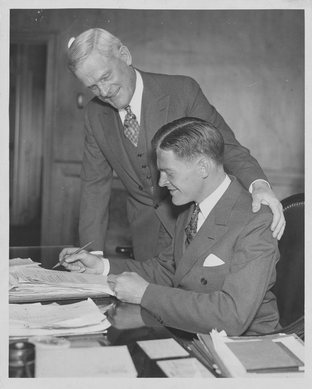 Black-and-white formal photograph of Thomas E. Wilson and Edward Foss Wilson. Edward is seated at a desk looking at papers. Thomas is standing with one arm on Edward's shoulders, looking at the same document as Edward. They are both white men wearing suits.
