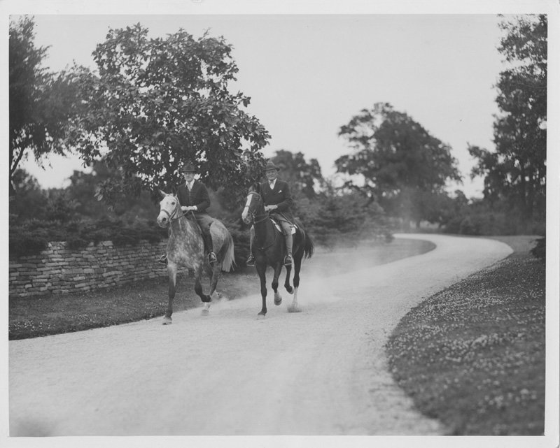 Black-and-white photograph of Thomas E. Wilson and Edward Foss Wilson riding horses down a curved gravel road. Trees are visible on both sides of the road, and a low stone wall lines one side of the road. Edward rides a dappled horse, and Thomas rides a dark horse. Both are white men wearing dark suits and fedora hats.