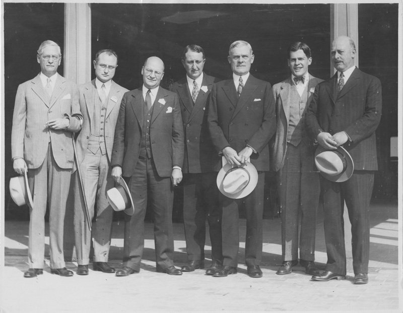 Black-and-white group photograph of seven white men standing outdoors in front of building.