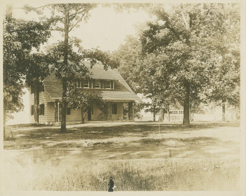 Black-and-white photograph of modest two-story home set amongst trees.