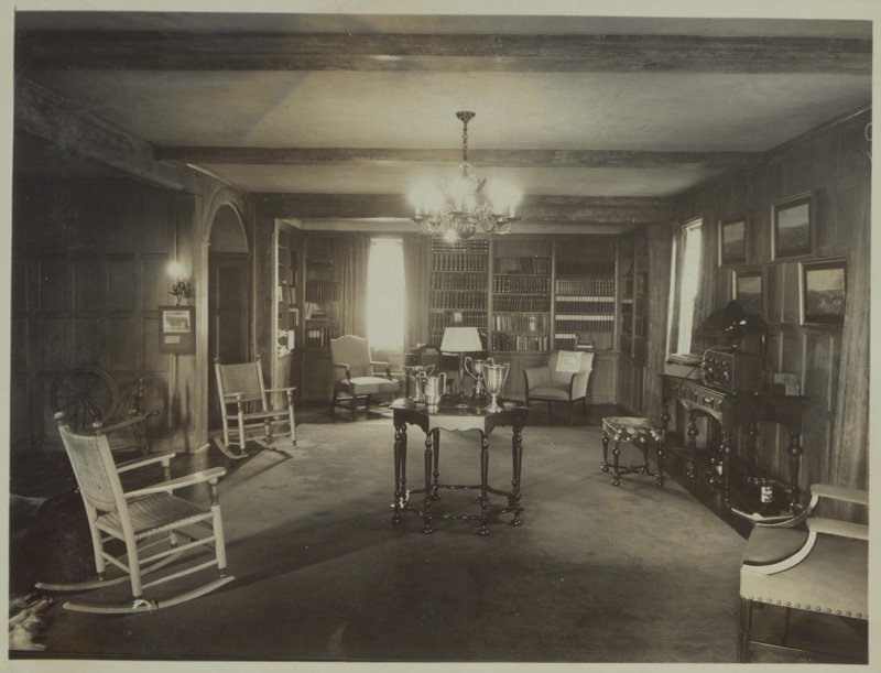 Black-and-white photograph of interior room of Edellyn Farm house. The library is a dark, wood-paneled room. A large area rug covers the floor. The ceiling has exposed beams. One wall is lined with bookshelves. Five chairs are set around the perimeter of the room. An octagonal wooden table with spindle legs is set in the center of the room. On it rests four trophies. A spinning wheel sets in a recessed part of the room. Another wall of the room has a rectangular table against it on which rests a radio. Four framed pictures are hung on the same wall above the table.
