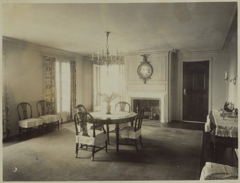 Black-and-white photograph of interior room of Edellyn Farm house. The dining room walls are painted a light color and a large area rug covers the floor. A circular wood dining table rests in the center of the room, with four wooden chairs with rounded backs around it. Two more dining chairs are set against one wall between two windows with floor-length drapes. A buffet table with a lace runner rests along the opposite wall. Another wall has a central fireplace, above which hangs an American Colonial-style mirror. Open, exterior french doors are to the left of the fireplace. A closed, interior wood door is to the right of the fireplace. A chandelier hangs above the dining table. A vase of flowers is in the center of the dining table.