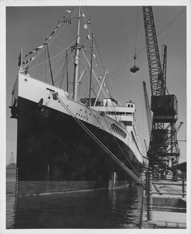 Black-and-white photograph of a large, docked ship. Cranes are hoisting barrels onto the ship.