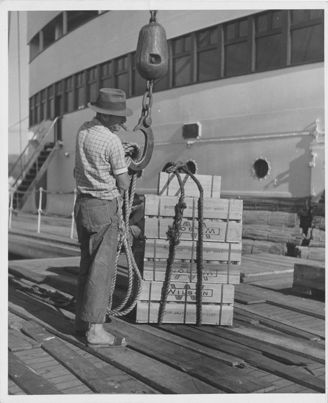 Black-and-white photograph of a man standing on a dock. He is attaching crates of Wilson products to a crane hook. He is wearing a hat and smoking a cigar.