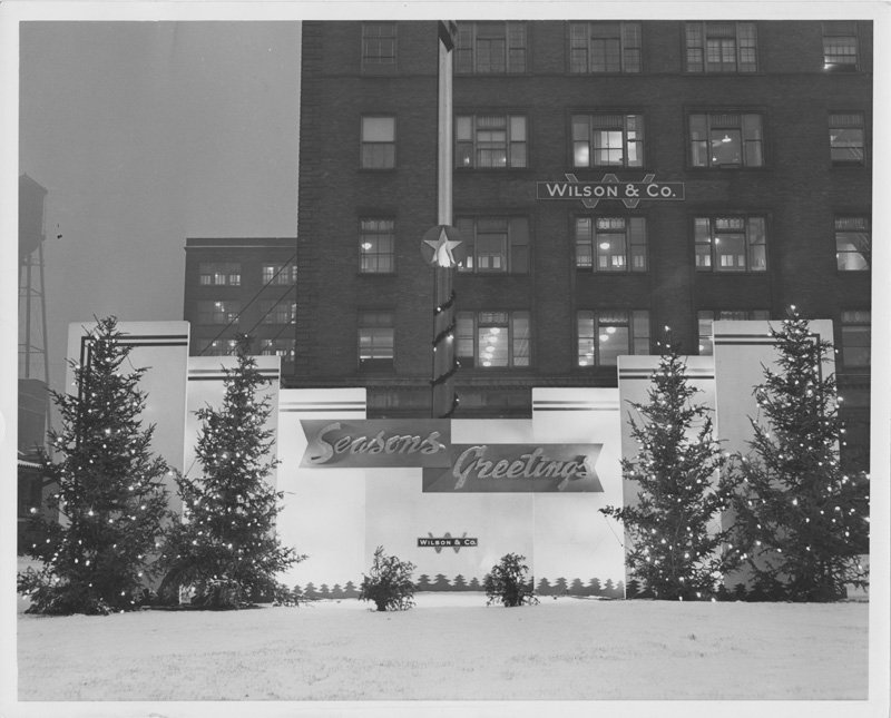 Black-and-white photograph of the exterior of a Wilson & Co. office building in the wintertime (presumably in Chicago). Snow is on the ground. A multi-story brick building is shown with a winter holiday display in front. Fir trees with lights strung on them flank a sign that reads "Seasons Greetings." The Wilson & Co. logo is below the sign. The Wilson & Co. sign is also on the brick building.