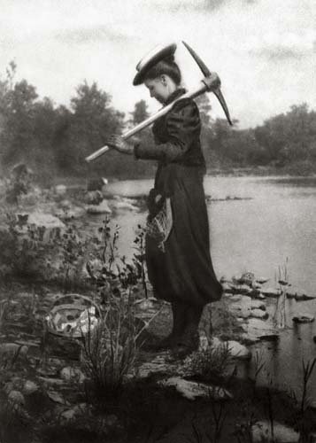 Photo of Zonia Baber in a swamp, carrying a pickax.